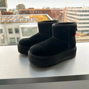 Svarta boots från UGG - Ett par svarta boots från UGG med en tjock sula och rund tå. Skorna är tillverkade i ett mjukt material och har en hög ankel.