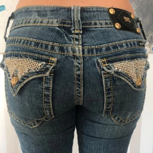 Blå jeans från Miss Me - Säljer ett par snygga blå jeans från Miss Me i storlek 28. De har en låg midja och en bootcut-stil som ger en cool look. Baktill finns det detaljerade fickor med glittriga paljetter och guldknappar som verkligen sticker ut. Perfekta för både vardag och fest!