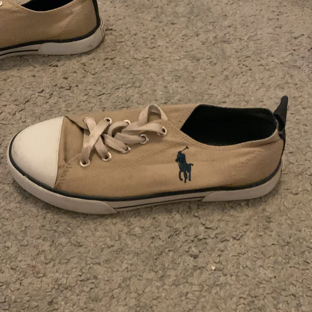 Säljer ett par snygga beige sneakers från Polo Ralph Lauren. Skorna har en vit tåhätta och vita snören. De är dekorerade med den klassiska Polo-loggan på sidan och har en svart sula med mönster. Perfekta för vardagsbruk och ger en stilren look.. Skor.