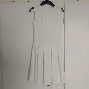 Det är en klänning som är vit. Den är från Nelly. Tygget är konstmaterial och har en dold kjeda i ryggen.