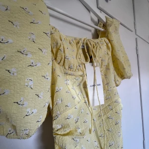 Adoore klänning  - En gul Adoore klänning med små vita blommor på. Fluffiga ärmar och ett knyte vid brösten. Går att justera längd genom att knyta snöre. Dragkedja i sidan.  ALDRIG ANVÄND - lappar på!!!  Pris går att diskutera. 