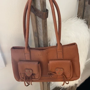 Handväska/axelväska från Morgan De Toi  - Handväska/axelväska från Morgan de toi. Förslutning dragkedja. 2 små ytterfack och 4 små innerfack i olika storlekar, en har dragkedja. Väskan är inte  2 färgad, utan det är ett fönster bredvid som reflekterar ljuset! Kan ta fler bilder vid intresse!