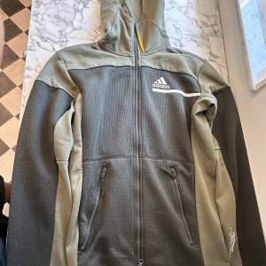 En militärgrön Adidas zip hoodie. Hoodien är luftig och sportig. Kan användas både för träning men även för att använda ute. Den har flera fickor för förvaring av små saker samt en stor luva. Hoodien har endast använts 5-10 gånger och är i gott skick