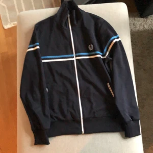 Svart kofta från Fred Perry - En svart kofta från Fred Perry med dragkedja och vita samt blå ränder. Koftan har en broderad logotyp på bröstet och är i storlek M. Koftan är i jättebra skick.