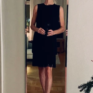 Svart festklänning, vintage, stlk 38, märke Martinette. - Svart vintageklänning för festsammanhang, märke Martinette. Ärmlös, klassisk 60-talsmodell med två dekorativa rader av fransar i kjollängden. Plagget är fodrat med ett svart fodertyg. Dragkedja bak. I nyskick. Stlk 38. Pris 250 kr.  FRI FRAKT.