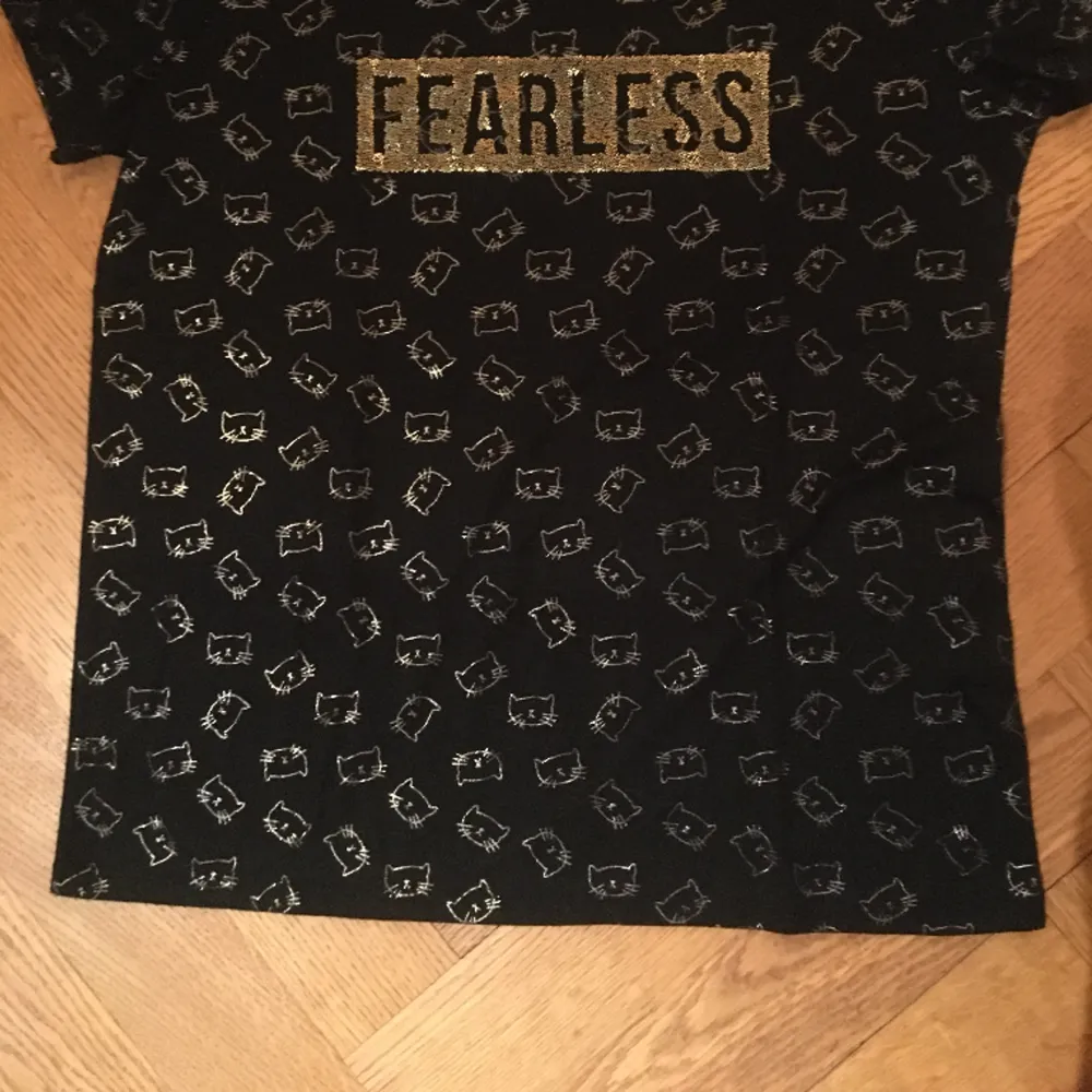 Svart t-shirt i fint skick med guldtext Fearless och kattansikte som liknar Hello Kitty, stl s. Pris: 40:-. T-shirts.