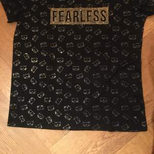 Svart t-shirt i fint skick med guldtext Fearless och kattansikte som liknar Hello Kitty, stl s. Pris: 40:-
