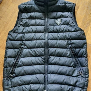 EA7 väst - Svart lätt dunväst från Emporio Armani. En knappt synlig lite reva på ena sidan annars perfekt skick. Inköpt 2022, sparsamt använd. Nypris ca 1400 kr. 