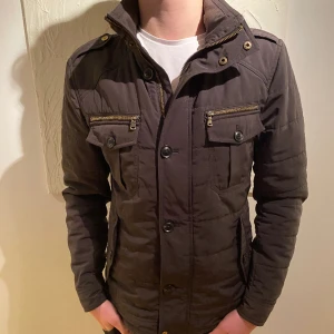 Field jacket - Säljer nu denna Field jacket från Riley, skick 8/10 då dragkedjan inte funkar men knapparna funkar lika bra, färgen är brun/mörkgrå! Model är ca 180