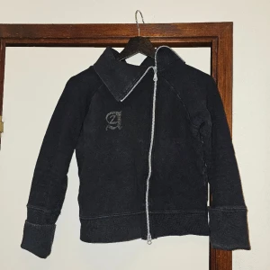 Långärmad tröja med asymmetrisk dragkedja - Beskrivning: Svart asymmetrisk tröja med tryck på rygg och bröst.  Märke: Storlek: M Skick: I gott skick, inte urtvättad som det ser ut som på bilderna, utan svart. Material:  Nakenkatt finns i hemmet