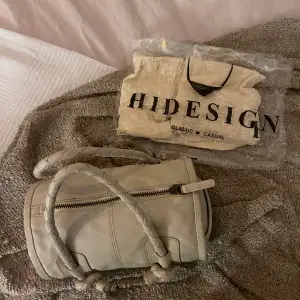 Fin liten väska av märket Hidesign.  Väskan är unikt cylinder formad och är en blandning av beige, och grå. Väskan är av äkta läder. Väldigt fint skick, aldrig använd. Dustbag medföljer!  För fler bilder eller frågor skriv till mig! ✨  