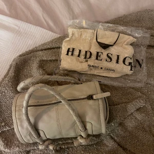 Stilren liten väska  - Fin liten väska av märket Hidesign.  Väskan är unikt cylinder formad och är en blandning av beige, och grå. Väskan är av äkta läder. Väldigt fint skick, aldrig använd. Dustbag medföljer!  För fler bilder eller frågor skriv till mig! ✨  