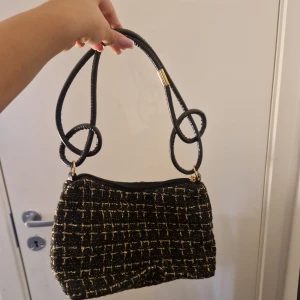 Handväska - Cool handväska i tyg. Svart med guldglitter typ. Bandet är av läder. Aldrig använd så i bra skick, dragkjedan funkar utan problem. Har aldrig haft användning för den så därför säljs den. Kommer tyvärr inte ihåg vart den är ifrån☺️