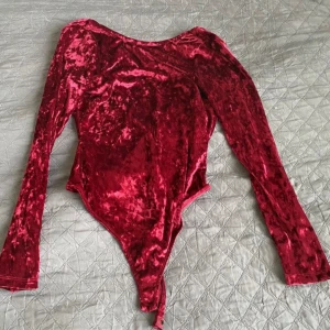 Röd sammetstopp - En röd långärmad sammetstopp med djup U-ringning i ryggen och bodysuit-design. Toppen har en glansig yta och är tillverkad i ett mjukt och stretchigt material.