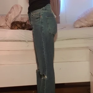 Blå jeansbyxor med hål - Ett par blå jeansbyxor med hål vid knäna. Byxorna har en hög midja och en rak passform. De är tillverkade i denim och har en klassisk femficksdesign. Köpta på H&M. Säljer pga att de är för stora. Storlek 36. Skriv för mer bilder.