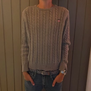 Grå stickad tröja från Maroy Classic - En grå stickad tröja från Maroy Classic med ett kabelstickat mönster. Tröjan har långa ärmar och en ribbstickad mudd vid ärmslut och nederkant. Märket har en broderad logotyp med en amerikansk flagga på bröstet.