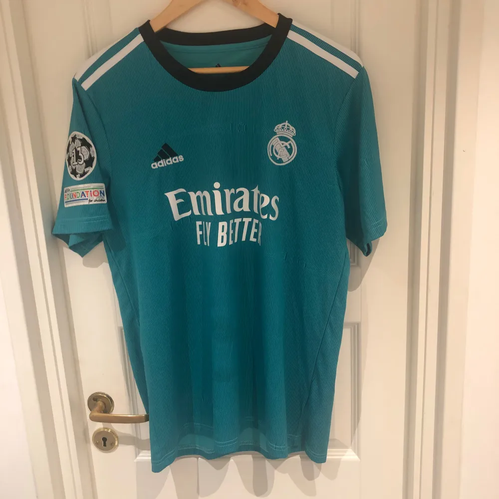 En blå fotbollströja från Adidas med Real Madrids logotyp och sponsortryck 'Emirates Fly Better' på framsidan. Tröjan har korta ärmar och ett svart rundhalsat krage. På baksidan finns texten 'VINI JR.' och nummer 20 i vitt. Priset Kan Alltid ändras. T-shirts.