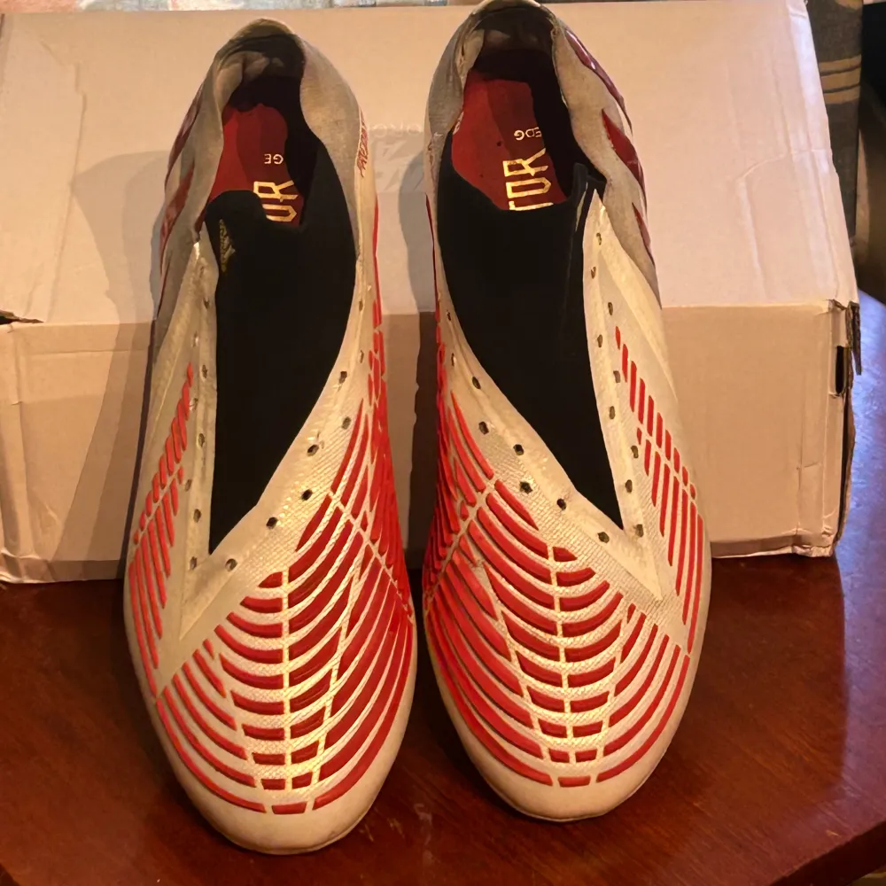 Till salu: Adidas Predator Edge i vitt och rött. Dessa skor är designade för ultimat prestanda på fotbollsplanen med en perfekt kombination av komfort, grepp och bollkontroll. Perfekt för spelare som vill ha överlägsen känsla och precision. . Skor.
