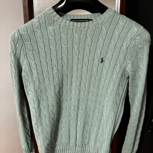 Grön stickad tröja från Ralph Lauren - En stilren och bekväm grön stickad tröja från Ralph Lauren med ett kabelstickat mönster. Tröjan har långa ärmar och ribbstickade muddar vid ärmslut och nederkant.