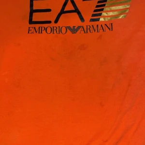 Orange t-shirt från Emporio Armani - En orange t-shirt från Emporio Armani med svart och guld text på framsidan.