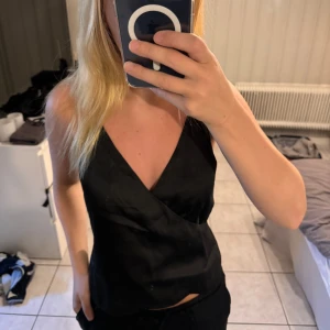 Svart V-ringad Satin Singlet från NA-KD - En elegant svart V-ringad satin singlet från NA-KD. Toppen har smala axelband och en figurnära passform, perfekt för festliga tillfällen.