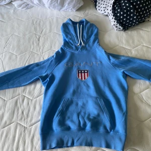 Blå hoodie från GANT - En blå hoodie från GANT med en broderad logotyp och ett sköldmärke på framsidan. Hoodien har en känguruficka och vita dragsnören vid huvan. Den är i storlek S och har en liten fläck på ärmen.