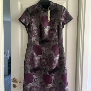 Mönstrad klänning - En elegant mönstrad klänning med korta ärmar. Klänningen har en blandning av mörka och ljusa färgtoner, inklusive vinröd och grå. Den är tillverkad av ett glansigt material och har en figurnära passform.