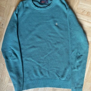Grön stickad tröja från Morris - En oliv grön stickad tröja från Morris med rund halsringning. Tröjan har långa ärmar och en liten broderad detalj på bröstet. Skicket på tröjan är ca en 9/10.