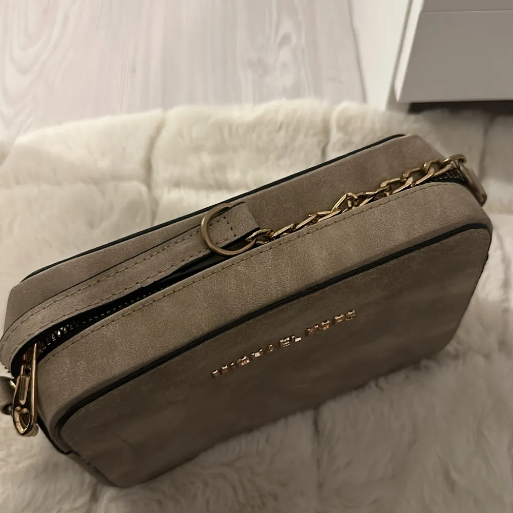 En elegant beige axelväska från Michael Kors med guldfärgade detaljer och en justerbar axelrem. Väskan har en dragkedja för säker förvaring. En liten streck på väskan men inget man tänker på.. Väskor.