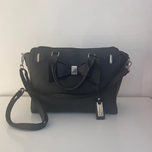 Svart väska - Svart stor väska med silvriga detaljer och en stor rosett på framsidan. Det följer med ett band till väskan som är justerbart. Väskan rymmer mycket och har tre innerfack varav ett har dragkedja, det finns även ett fack på baksidan.  Märket är Catwalk