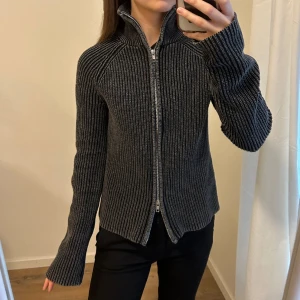 Grå cardigan - Grå tröja i ribbat med mycket stretch och tvåvägsdragkedja. Kragen är hög men går att vika ner. Långa mysiga ärmar.
