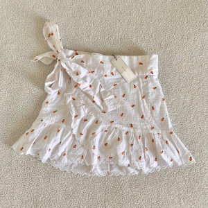 Neo Noir vit omlottkjol med apelsiner - Otroligt söt kjol från Neo Noir med apelsiner. Omlottkjol som knyts med rosett på sidan. Ny med tags kvar, utan anmärkningar.