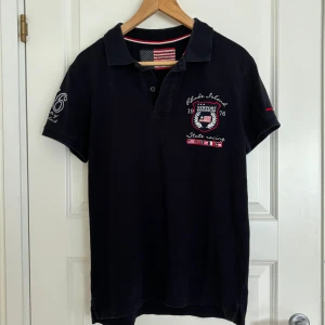 Newport piké - Detta är min brors mörkblå piké från märket Newport. Han har vuxit ur den och använder den därför inte längre. Den har ett flertal tryck; dels på respektive arm, dels på bröstet och ryggen. Den är i gott skick utan några defekter.