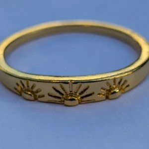 Guldfärgad ring med solmotiv - En elegant guldfärgad ring med ingraverade solmotiv. Ringen har en slät yta och är dekorerad med flera små solsymboler runt om.