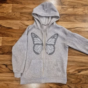 Grå huvtröja med fjärilsmotiv - Oversized fit: En grå huvtröja med dragkedja och ett stort fjärilsmotiv på framsidan. Tröjan har långa ärmar och två fickor framtill. 