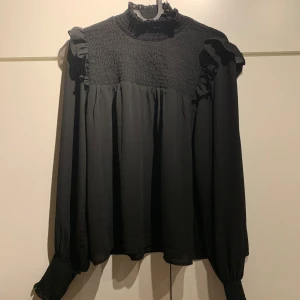 Svart blus - En svart blus med polo, volanger och smock från BikBok. Använd endast en gång så nyskick. Pris kan diskuteras!