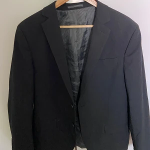 Svart klassisk kavaj - En stilren svart kavaj med två knappar framtill och klassiska slag. Fodret har diskreta ränder för en snygg touch. Passar perfekt till skjorta och byxor för en clean look. Enkel bröstficka och två fickor framtill.