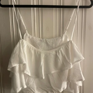Vit offshoulder volangtopp Stradivarius - Superfin vit offshoulder topp från Stradivarius med tunna axelband och dubbla volanger. Toppens smockade liv ger en snygg passform och den är tillverkad i ett lätt, luftigt tyg som känns perfekt för varma dagar. Stilren och trendig look!