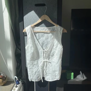 Vit broderad väst med knyt - Perfekt då den har inga fläckar eller missfärgningar! vit väst med broderade blommor och blad, v-ringad och knyt framtill. Västen är i ett lätt material, perfekt för varmare dagar. Snygg att styla över en t-shirt eller klänning för en romantisk vibe.
