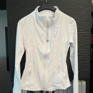 Vit Lululemon nulu-jacka strl 2 - Snygg vit nulu-jacka från Lululemon med figurnära passform, hög krage och dragkedja framtill. Jackan har långa ärmar, två fickor med dragkedja och är gjord i ett stretchigt syntetmaterial som andas. Perfekt för dig som gillar sportig och clean stil. Strl 2 - utan etikett, men ej använd, passade ej bra 