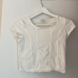 Vit croppad korsett-topp - Säljer en vit croppad korsett-topp från Bik Bok med korta ärmar och markerade sömmar framtill som ger en snygg siluett. Toppen har rund halsringning och är gjord i mjuk bomull, perfekt till jeans eller kjol.