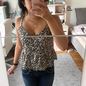 Blommig linnetopp från Gina Tricot - Supersöt blommig linnetopp från Gina Tricot i svart med vita och beige blommor. Toppen har smala axelband, knappar framtill och en volangkant nedtill som ger en extra feminin touch. Ge ett valfritt pris!!