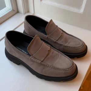 Garment Project Phil Loafer – Ardesia Suede - Helt nya och oanvända loafers från Garment Project – modell Phil Loafer i exklusiv ardesiafärgad mocka. Klassisk slip-on-design med moc-tå och rejäl, chunky gummisula. Tillverkade i Portugal med antibakteriell innersula för extra komfort. En stilren sko som passar både till vardags och mer uppklädda tillfällen. Nypris i butik: ca 2000 kr.