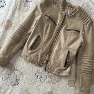 Beige skinnjacka från Livelo, stl 36 - Snygg beige skinnjacka från Livelo med coola detaljer som ribbade partier på axlar och ärmar, fyra dragkedjefickor och spännen bak. Jackan har en klassisk bikerstil och stängs med dragkedja framtill. Perfekt för dig som vill ha en stilren och edgy look.