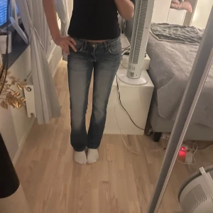 Lågmidjade Blå bootcut jeans  - Snygga lågmidjade blå bootcut jeans med slitningar och broderade detaljer på bakfickan. Jeansen har klassisk femficksmodell, låg midja och råa kanter nertill. Perfekta för dig som gillar en avslappnad och trendig look.