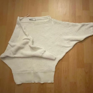 Vit ribbad stickad tröja från NAKD - Vit stickad tröja från NAKD med ribbad struktur och bred mudd nertill. Tröjan har lång ärm och en lös, oversized passform som ger en chill och trendig look. Perfekt att slänga på sig när det är lite kyligt ute.