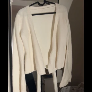 Vit stickad kofta med knytband - Mysig vit stickad kofta med långa ärmar och knytband i midjan. Perfekt att slänga över en topp för en chill och stilren look. Koftan har en öppen front och ribbade muddar vid ärmslut och nederkant.