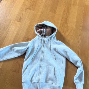 Grå hoodie från Burberry med dragkedja - Snygg ljusgrå hoodie från Burberry med klassiskt rutigt foder i huvan. Tröjan har dragkedja framtill, snörning vid huvan och två stora fickor. Perfekt för dig som gillar stilrena och exklusiva plagg med en twist.