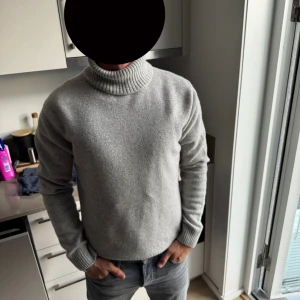 Ljusgrå polotröja från Oscar Jacobsson  - Säljer en stilren ljusgrå polotröja i mjuk ull/kashmir mix. Tröjan har en hög, ribbad polokrage och långa ärmar med ribbade muddar. Perfekt för dig som gillar en clean och enkel look med en touch av klass.