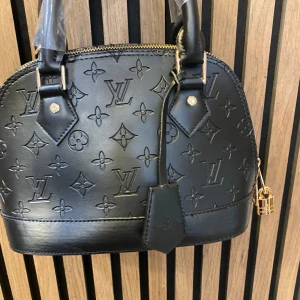 Svart  handväska i skinn - Säljer en svart handväska  i skinn . Väskan har rundad form, två handtag och guldfärgade metalldetaljer samt dragkedja och hänglås. Perfekt för dig som gillar lyxiga accessoarer.